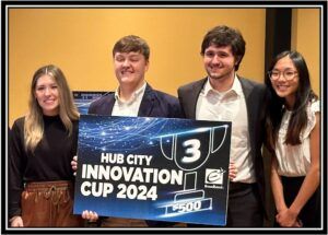 Four smiling young adults stand indoors holding a large sign that reads Hub City Innovation Cup 2024, 3rd, $500, Broadcom. They appear to be at an event or competition.