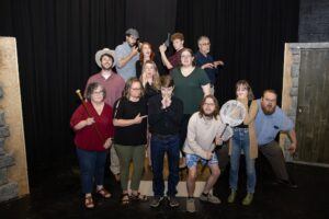 A group of thirteen people pose on a stage in dramatic and playful stances, with black curtains behind them. One person holds a staff, another a butterfly net. Their expressions and gestures vary from serious to silly.