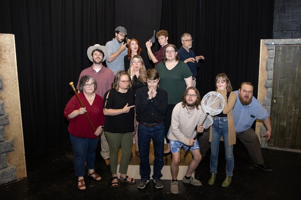 A group of thirteen people pose on a stage in dramatic and playful stances, with black curtains behind them. One person holds a staff, another a butterfly net. Their expressions and gestures vary from serious to silly.