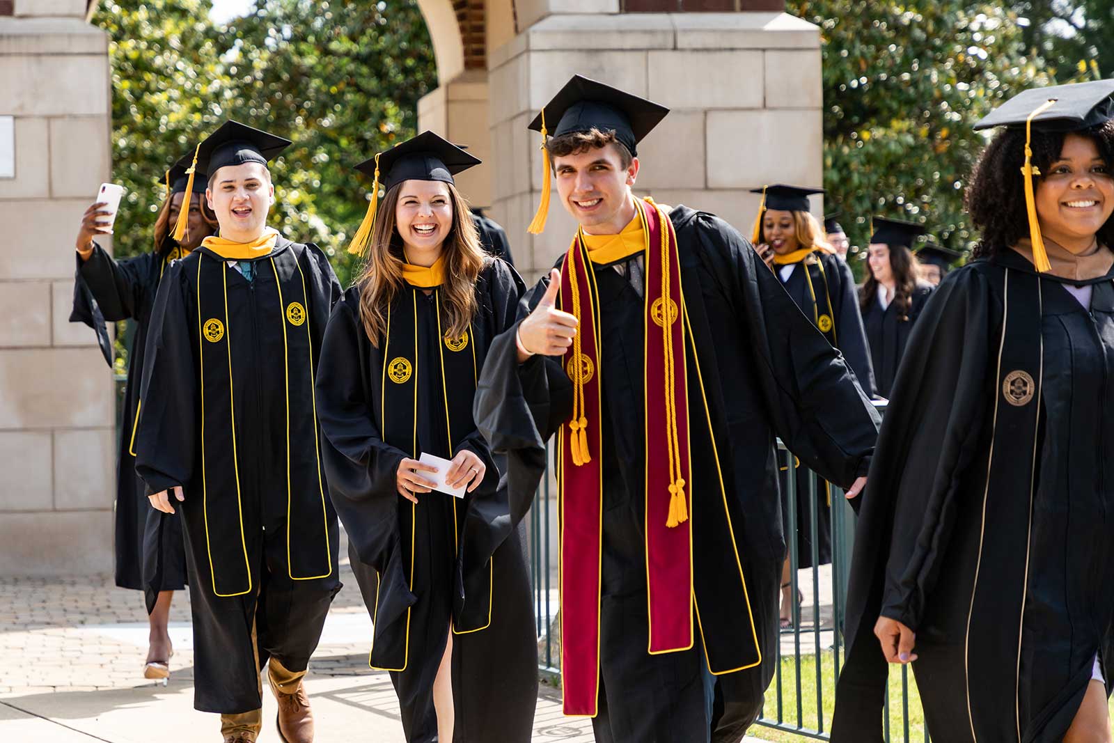 students in regalia outside walking into commencement giving a thumbs up