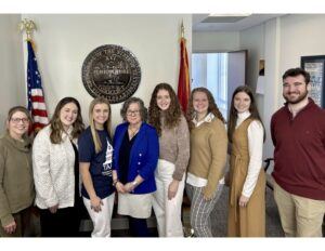 Seven people standing in a row, smiling in an office with flags and a large seal for the Department of Agriculture on the wall behind them. Four women are on the left, two women and one man are on the right.