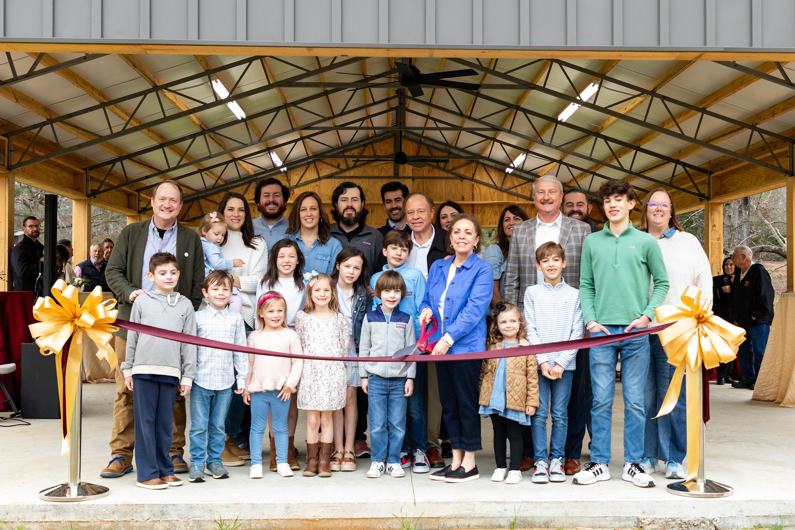 A large group of adults and children stand together in a covered outdoor pavilion, smiling for a photo behind a red ribbon with gold bows, suggesting a ribbon-cutting ceremony.