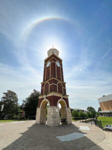 A tall brick clock tower stands under a bright sky, with a circular halo around the sun visible above it. The surrounding area features trees, a curved sidewalk, and a building to the right.
