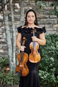 A woman in a black off-the-shoulder dress stands outdoors in front of a stone wall and greenery, holding a violin in one hand and a viola in the other.