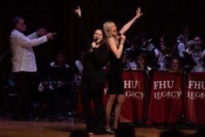 Two women in black outfits stand back to back on stage, singing passionately into microphones with one arm raised. A band plays behind them, with FHU Legacy banners visible on music stands. A conductor stands to the left.