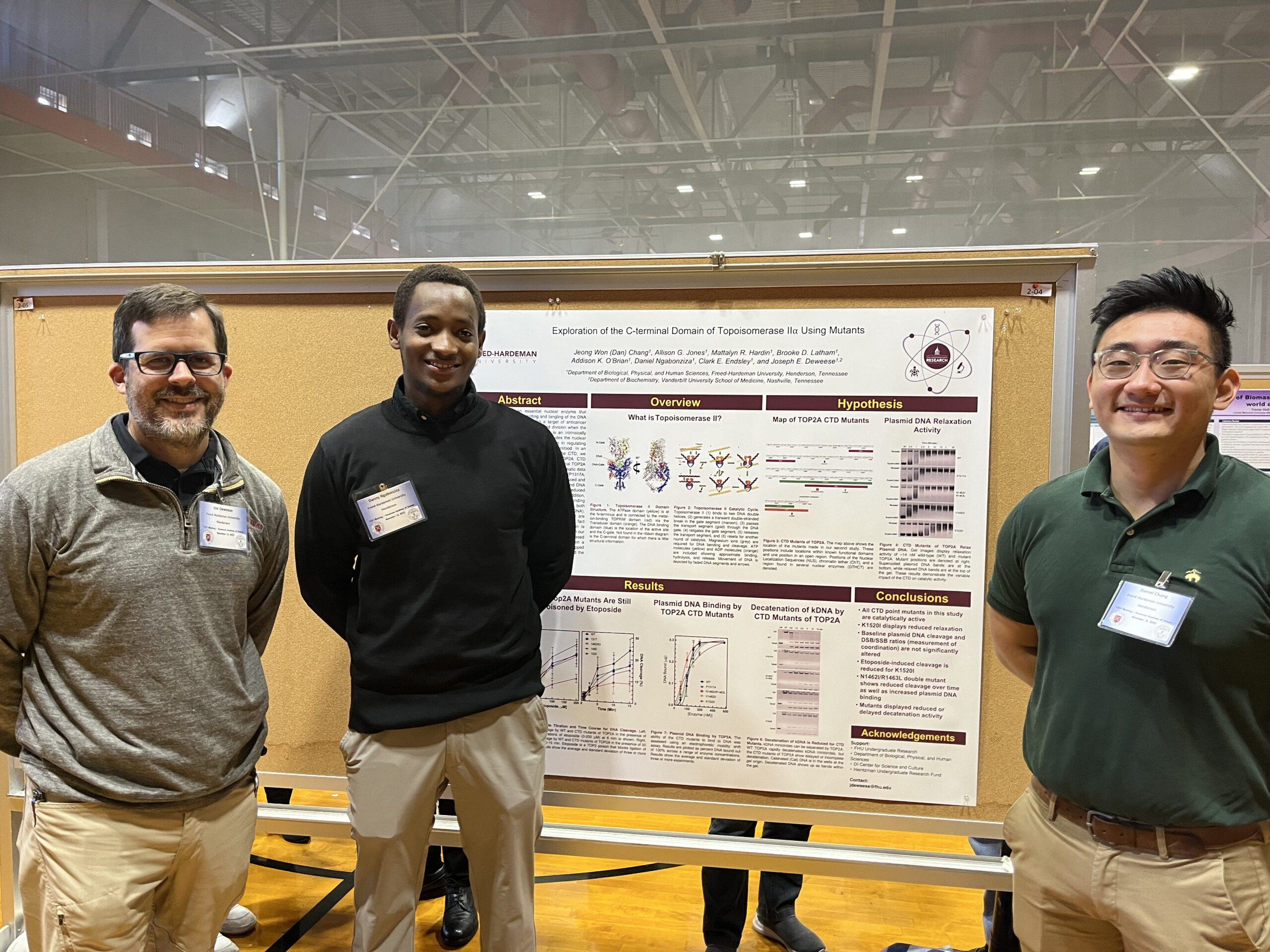 Three people stand smiling in front of a scientific research poster displayed on a corkboard in a gymnasium. The poster features text, charts, and diagrams about Toxoplasma research.