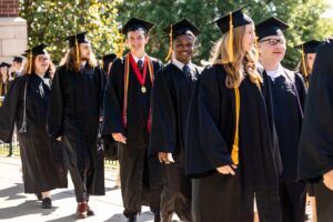 A group of graduates in black caps and gowns walk together outdoors on a sunny day, smiling and chatting, with trees and a building in the background.
