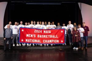 A mens basketball team poses on stage, smiling and holding a large red and blue banner that reads 2024 NAIA Mens Basketball National Champion. Coaches and staff stand at both ends of the group.