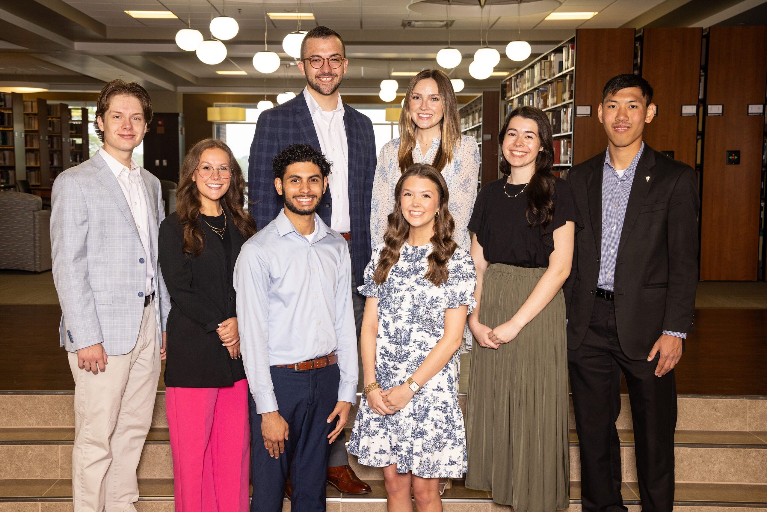 Eight young adults, four men and four women, stand in two rows on indoor steps, dressed in semi-formal attire, smiling at the camera. The background shows bookshelves and round lights in a library setting.
