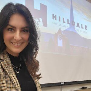 A woman with long dark hair and a plaid blazer smiles in front of a projected screen displaying Hilldale Christian Child Care Center and an image of a church building with a tall steeple.