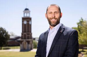 A man in a checked suit jacket and light dress shirt smiles outdoors, with a brick clock tower and green trees visible in the background under a clear sky.