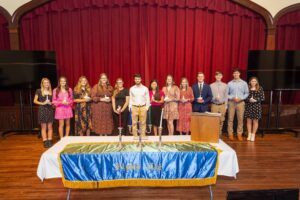 A group of thirteen young adults stand in a line on a stage with red curtains, holding awards. In front of them is a table covered with a blue and gold cloth that reads “Alpha Chi.”.
