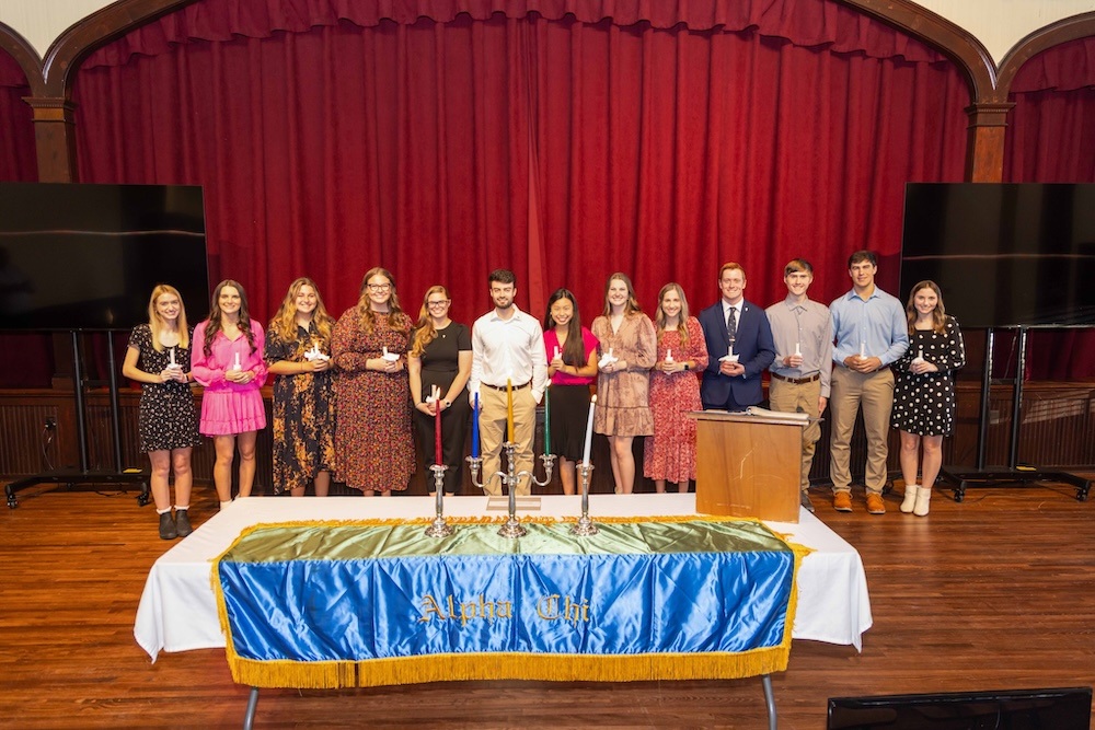 A group of thirteen young adults stand in a line on a stage with red curtains, holding awards. In front of them is a table covered with a blue and gold cloth that reads “Alpha Chi.”.