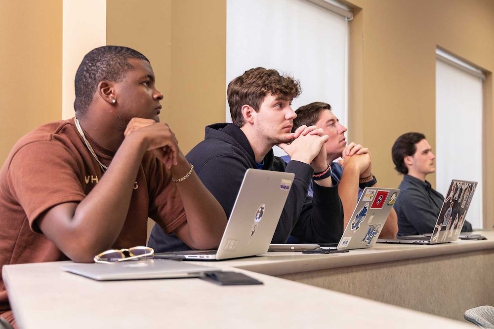 Four students sit at desks in a classroom, each with a laptop, attentively listening. They rest their chins on their hands and face forward. The background shows light-colored walls and windows with shades.