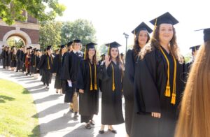Smiling graduates standing in a line, with several waving at the camera. The Freed-Hardeman Belltower is in the background