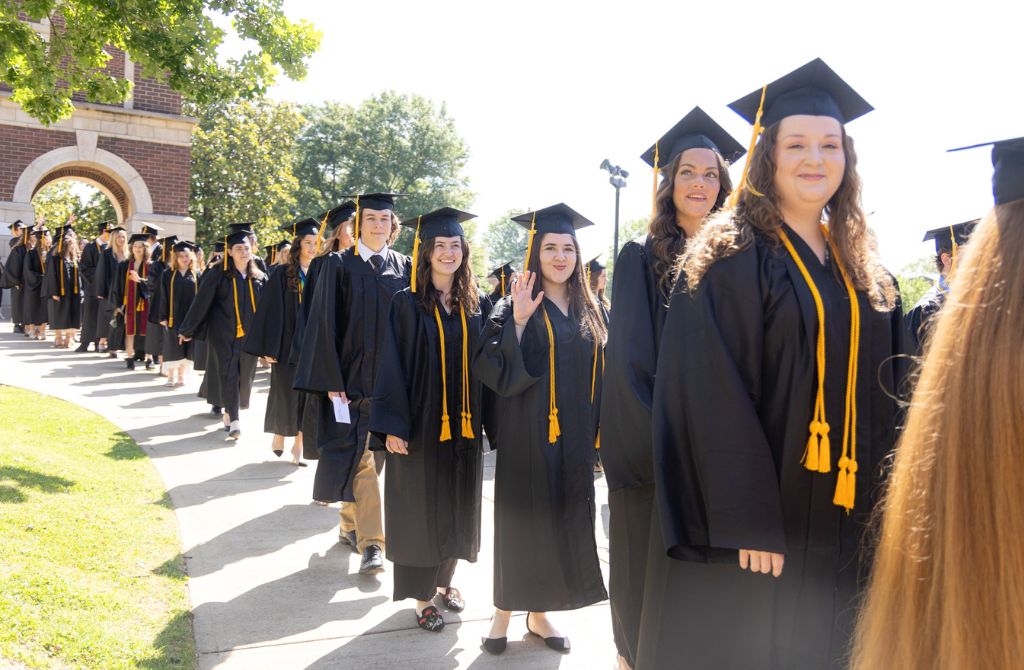 Smiling graduates standing in a line, with several waving at the camera. The Freed-Hardeman Belltower is in the background