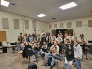 A group of people in a music classroom, many wearing matching vests with patches, pose with musical instruments, including guitars and brass. Some are seated, some standing, all smiling at the camera. Music stands and chairs fill the room.