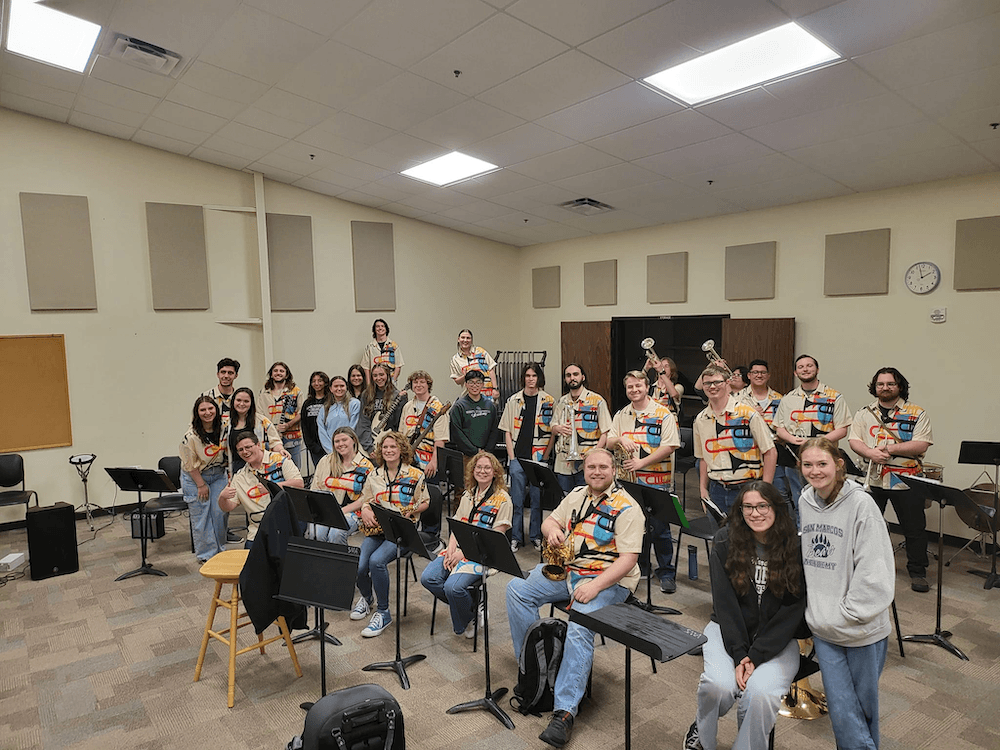 A group of people in a music classroom, many wearing matching vests with patches, pose with musical instruments, including guitars and brass. Some are seated, some standing, all smiling at the camera. Music stands and chairs fill the room.