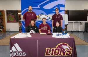 A high school baseball player sits at a table with coaches and family, smiling for a signing ceremony. The table displays two jerseys, hats, and a Lions logo, with a large eagle banner in the background.