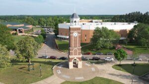 Aerial view of a red-brick clock tower with a white roof, surrounded by a circular pathway, trees, parked cars, and large buildings in the background on a sunny day.