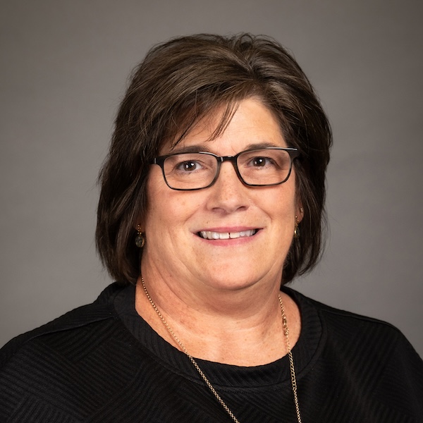 A woman with short brown hair and glasses smiles at the camera. She is wearing a white shirt with a dark plaid pattern and small earrings, set against a plain dark background.
