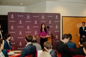 A woman stands at a podium speaking to an audience in a conference room. Behind her is a maroon Freed-Hardeman University backdrop. Several people are seated and listening, and a man stands near a door in the background.