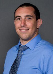 A man with short dark hair and a trimmed beard smiles at the camera. He is wearing a light blue dress shirt and a gray patterned tie, posed against a plain gray background.