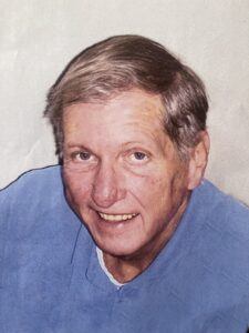A smiling older man with short, light brown hair wearing a light blue collared shirt, looking slightly up and to the side against a plain, light background.