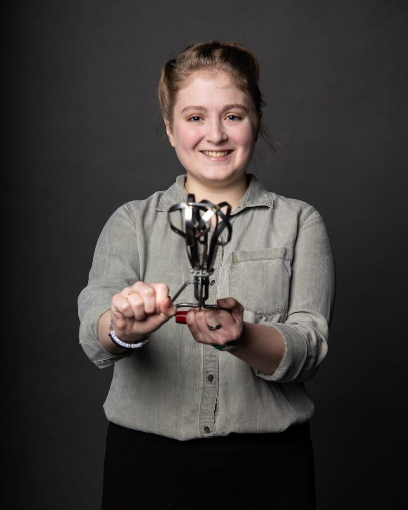 A smiling person in a grey button-up shirt holds a small metal trophy or mechanical object toward the camera, standing against a plain dark background.