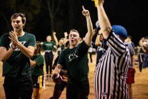 A flag football player celebrates with a football in hand and finger raised as teammates cheer in the background. A referee in a striped shirt holds up a yellow flag on a nighttime field.