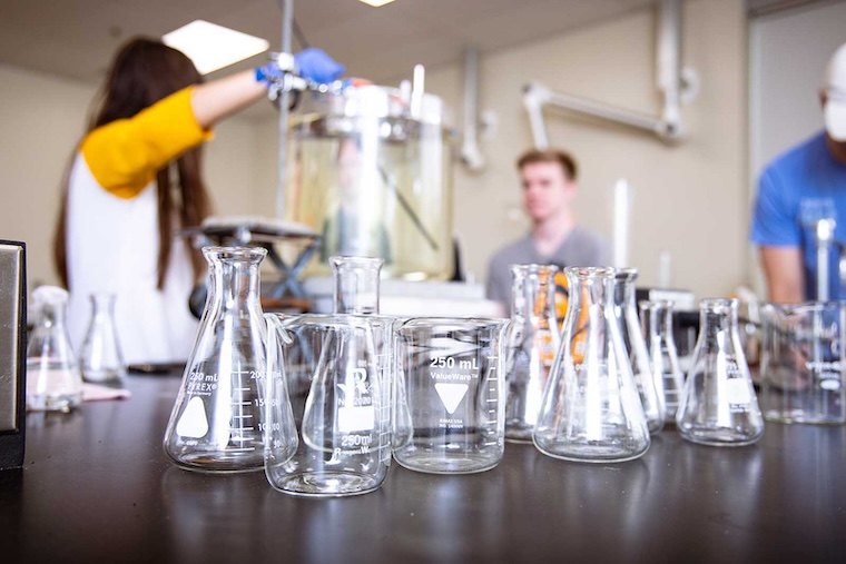 Several empty glass beakers and flasks are arranged on a black laboratory table; in the background, people in casual clothes work with lab equipment in a bright, modern science lab.