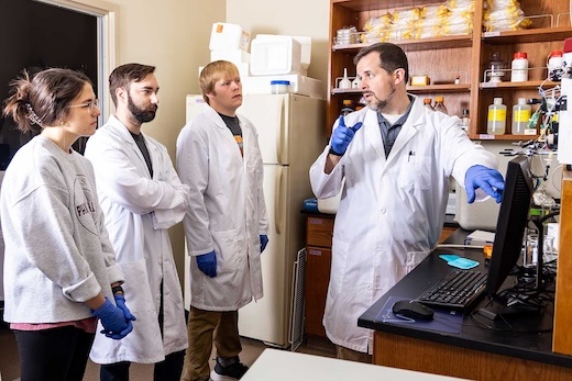 A scientist in a lab coat and gloves gestures while talking to three young adults, also in lab coats and gloves, in a laboratory with shelves holding bottles and equipment.