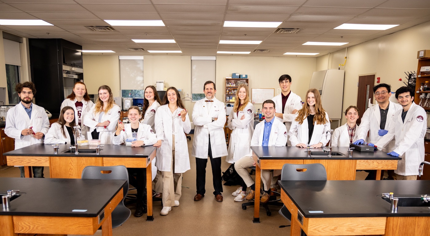 A group of students in lab coats smile alongside their professor in one of the labs on campus
