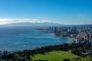 Aerial view of a coastal city with high-rise buildings along the shoreline, turquoise ocean, green park area in the foreground, and distant mountains under a clear blue sky.