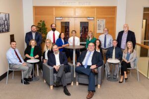 A group of fifteen professionally dressed adults poses for a group photo in a modern office lobby. Three people are seated in front, with the rest standing and sitting around two small tables under a sign that reads Carter Suite.