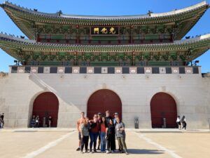 A group of seven people poses in front of the grand, ornate main gate of a traditional Korean palace with three large red doors and intricate green and gold detailing under a clear blue sky.