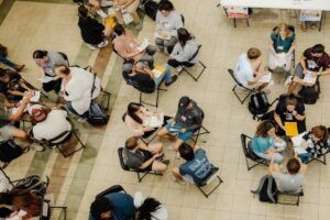 A group of people sit in pairs on chairs arranged in rows on a tiled floor, engaged in conversations or activities, with papers and folders in hand. The scene appears to be a workshop or group event viewed from above.