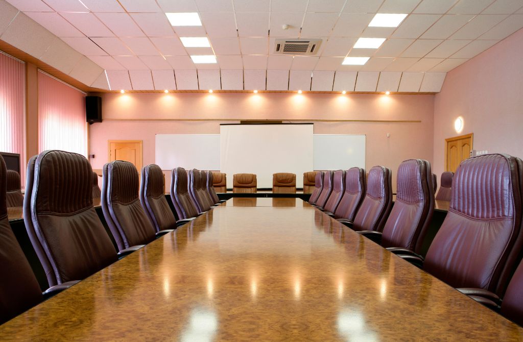 An empty office room with brown leather seats pulled around a brown glossy table. It is well lit.