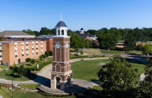 A tall brick clock tower stands in a grassy open area, surrounded by paths, trees, and a multi-story red-brick building under a clear blue sky. Other campus buildings are visible in the background.