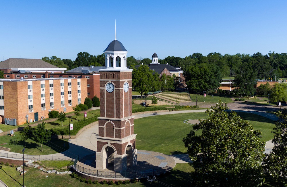 A tall brick clock tower stands in a grassy open area, surrounded by paths, trees, and a multi-story red-brick building under a clear blue sky. Other campus buildings are visible in the background.