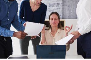 A woman sits at a desk with her eyes closed and hands in a meditative pose, while three colleagues around her hold out papers, creating a busy office scene.