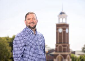 A man wearing a blue plaid shirt smiles while standing outdoors with a clock tower in the blurred background and trees on a sunny day.