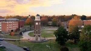 Aerial view of a campus clock tower surrounded by a circular walkway, grassy area, and trees with autumn foliage. Brick buildings and cars are visible under a colorful sunset sky.