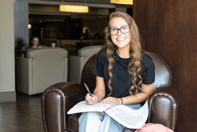 A young woman with long curly hair and glasses sits in a leather chair, smiling at the camera while holding a pen and writing in a notebook. She is casually dressed and appears to be studying in a lounge area.