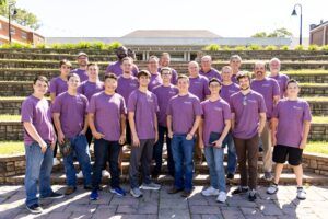 A group of about 23 people, mostly men of various ages, stand on stone steps outdoors, all wearing matching purple t-shirts and smiling at the camera. A building is visible in the background.