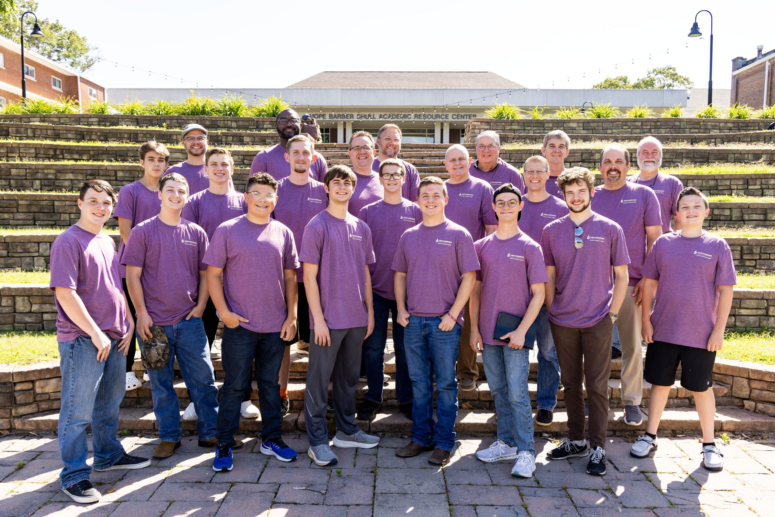 A group of about 23 people, mostly men of various ages, stand on stone steps outdoors, all wearing matching purple t-shirts and smiling at the camera. A building is visible in the background.