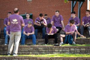 A group of young men wearing matching purple shirts sit on outdoor stone steps, listening to an instructor who stands facing them, also wearing a similar shirt. Several students are reading or taking notes.