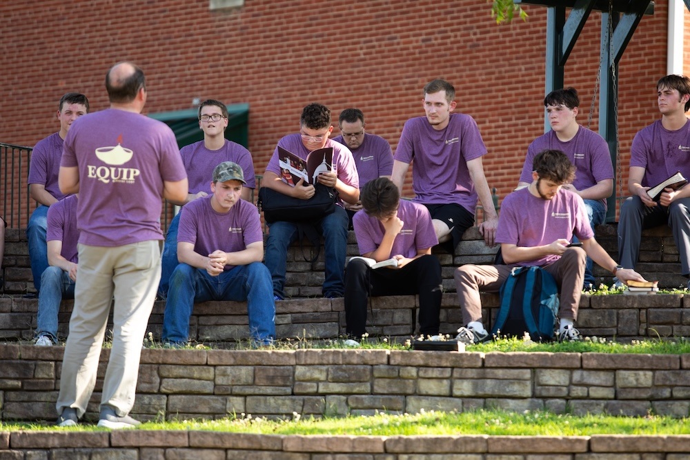 A group of young men wearing matching purple shirts sit on outdoor stone steps, listening to an instructor who stands facing them, also wearing a similar shirt. Several students are reading or taking notes.