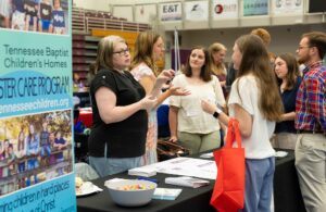 A group of people converse at a booth for Tennessee Baptist Children’s Homes during an indoor event. One woman holds a red tote bag while brochures, a bowl of candy, and posters are on the table.