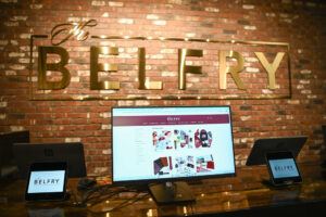 A computer monitor and two tablets on a counter display The Belfry’s website, with a large gold BELFRY sign on a brick wall in the background.